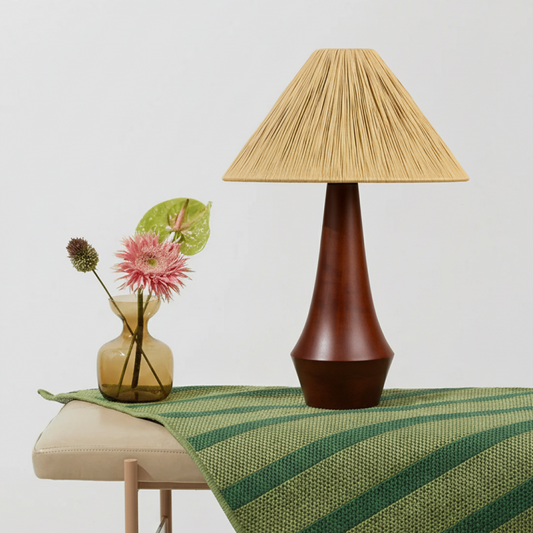 Table lamp with a woven shade on a green tablecloth next to a vase with flowers.