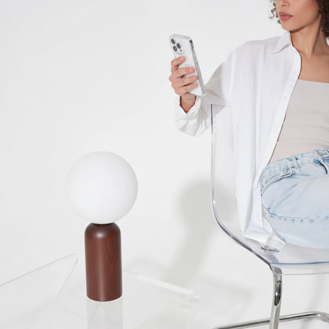 Person sitting on a clear chair holding a phone, with a white lamp in the foreground.